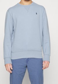 Světle modrý bavlněný svetr se zaobleným výstřihem, žebrovanými manžetami a lemem. Obsahuje malé námořnické logo na levé části hrudi.