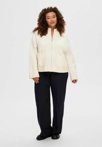 Crèmekleurige zip-up sweater met twee voorzakken, ribgebreide manchetten en zoom, en een zachte, gestructureerde stof, gecombineerd met donkere broeken.