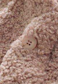 Puszysty beżowy materiał o miękkiej teksturze, z okrągłym, jasnym guzikiem z dwoma otworami przyszytym do tkaniny.