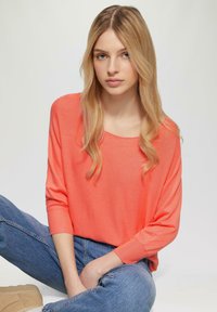 Korallfarbener Oversize-Pullover mit weitem Ausschnitt, strukturiertem Strick und Dreiviertelärmeln, kombiniert mit blauen, hoch taillierten Jeans.