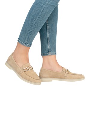 Beige suède loafers met een decoratieve kettingaccent, platte rubberen zool en ronde neus, gedragen met opgerolde blauwe spijkerbroeken.