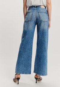 Pogled straga na osobu koja nosi visokostrukirane, široke plave traperice, bijelu majicu i crne sandale s petom na jednobojnoj pozadini.