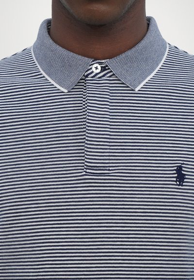 Polo de rayas azul marino y blanco con tapeta de dos botones, cuello plano y un pequeño logotipo azul marino en el lado izquierdo del pecho. Material de algodón.