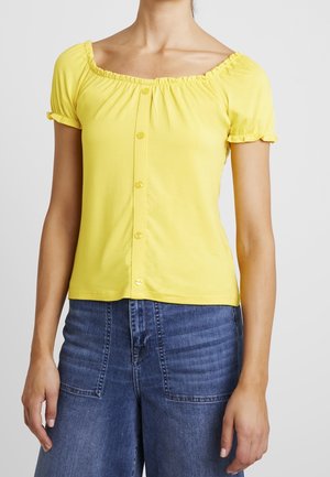 Corto top giallo a maniche corte con scollatura volant e chiusura a bottone davanti. Tessuto morbido e design aderente, abbinato a pantaloni in denim blu.