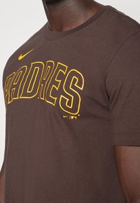 Brun bomull t-shirt med ordet "PADRES" i fet gula bokstäver, med en Nike-logotyp. Korta ärmar och standard rund halsringning.