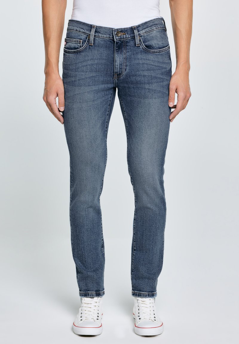 Mees, kes kannab klassikalise lõikega siniseid teksaseid ja valgeid tossusid, seisab heleda tausta ees, käed vabalt külgedel.