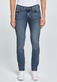 Mężczyzna w przylegających niebieskich dżinsach i białych trampkach, stojący na tle jednolitego jasnego tła, z rękami swobodnie opuszczonymi wzdłuż ciała.