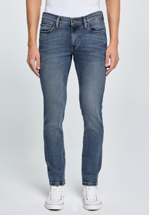 Férfi, aki slim-fit kék farmert és fehér tornacipőt visel, világos egyszerű háttér előtt áll, kezét nyugodtan oldalán tartva.