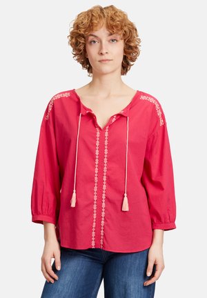 Blouse en coton rouge avec décolleté en V, manches trois-quarts, broderie décorative crème le long des épaules et au centre, liens réglables avec des franges.