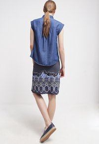 Camicia senza maniche in denim con orlo curvo, abbinata a una gonna a pieghe di lunghezza al ginocchio con motivi geometrici blu e scuri. Indossata con espadrillas blu.