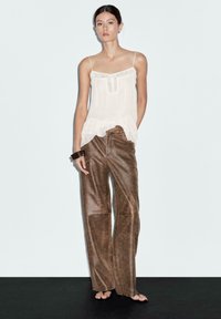 Woman wearing a white ruffled sleeveless top and wide-leg brown leather pants, accessorized with dark bangles, standing on a black floor.