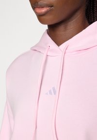 Hoodie rose clair en tissu doux avec une poche avant, des cordons de serrage ajustables et un logo Adidas argenté discret sur la poitrine.