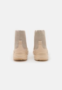 Sneaker in maglia beige a forma di calza viste da dietro, con tomaie testurizzate e suole in gomma spesse su uno sfondo bianco.