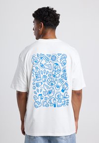White cotton t-shirt featuring a blue graphic design on the back, consisting of abstract shapes, eyes, hands, and various patterns.