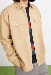 Beigen värinen napillinen puuvillapaita, jossa on kaksi rintataskua, painonapit ja väljä leikkaus, puettuna monivärisen raidallisen paidan päälle.