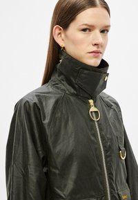 Vahatatud must jakk kõrge kraega, kuldsete detailide ja tõmblukuga, rinnakohal aasaga, matt-textuuriga.