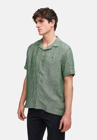 Camicia a maniche corte verde con bottoni, realizzata in tessuto testurizzato con leggere variazioni di colore e bottoni dall'aspetto legnoso, caratterizzata da un piccolo logo.
