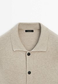 Beige gebreide cardigan met een geribde kraag, twee zwarte knopen en een gladde, zachte textuur. Label zichtbaar binnenin de kraag.