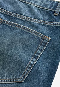 Jeans din denim cu o spălare albastră închisă, având cusături vizibile, patru buzunare și o țesătură texturată. Designul buzunarului din spate este unghiular.