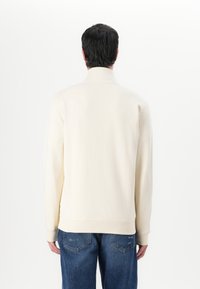 Felpa beige con collo alto, polsini e orlo a costine. Indossata con jeans in denim blu, vista da dietro su uno sfondo bianco.