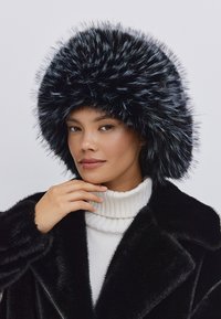 Sort faux fur hat med et voluminøst, fluffy design. Bæres med en hvid ribbet turtleneck under en sort frakk. Glat tekstur.