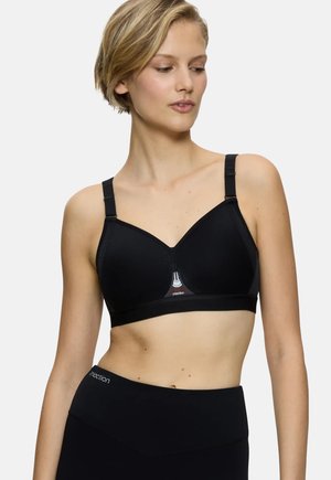 Femme portant un soutien-gorge de sport noir et un legging taille haute assorti, regardant légèrement vers le bas et sur le côté sur un fond uni.