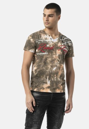 Junger Mann mit kurzen Haaren, der ein braunes Batik V-Ausschnitt T-Shirt mit rotem und weißem Text und schwarze Cargohosen mit Reißverschlussdetails trägt.