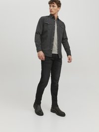 Camisa de botones gris oscuro con dos bolsillos en el pecho sobre una camiseta gris clara, combinada con jeans ajustados negros y botas negras hasta el tobillo.