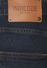 Donkerblauwe denim jeans met een tan leren merkpatch met verhoogde tekst, vastgezet met metalen klinknagels en contrasterende oranje stiksels.