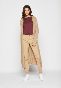 Lang beige fuzzy cardigan, burgunder topp og lyse khaki bukser med merkelapp. Hvite sneakers, glatt tekstur og avslappet passform.