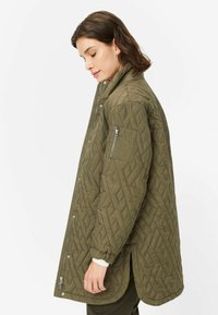 Gepolsterte olivgrüne Jacke mit hohem Kragen, seitlichen Schlitzen, Druckknöpfen und einer Reißverschlusstasche am linken Ärmel. Weiches, strukturiertes Material.