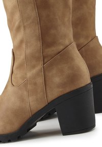 Brown faux leather knee-high boots with a block heel, featuring side cutouts, subtle texture, and stitching details.