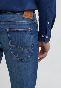 Siniset farkut, joissa on "Lee"-merkintäinen nahkapaikka vyötäröllä, takataskut ommeltuina ja suora leikkaus.