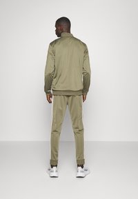 Olivovo zelený teplákový komplet so zipsovým kabátom a zodpovedajúcimi nohavicami, s bielymi bočnými pruhmi a elastickými manžetami. Hladká, ľahká tkanina.