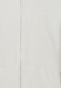 Hvit zip-up hettegenser med en myk tekstur, med frontglidelås, kengurulommer og en subtil logo med lys skrift på brystet.