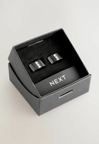 Schwarze Manschettenknöpfe mit geschwungenem Design, verziert mit einem klaren rechteckigen Steinakzent, präsentiert in einer Box mit weichem schwarzen Innenfutter und "NEXT"-Label.