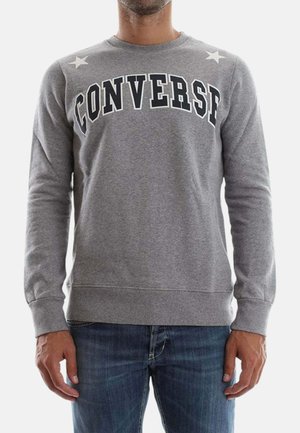 Uomo che indossa una felpa grigia a maniche lunghe con il testo "CONVERSE" e due stelle bianche vicino alle spalle, abbinata a jeans blu.