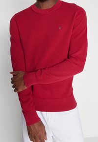 Suéter de punto ribeteado rojo con mangas largas, cuello redondo y pequeño logo en el pecho. Textura suave y diseño ajustado.