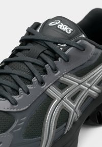 Gros plan d'une chaussure de running Asics noire et grise avec un mesh texturé, des lacets détaillés et des bandes logo argentées sur le côté.