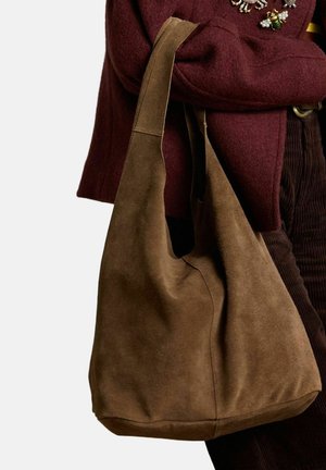 Bruine suède hobo-tas met een zachte textuur, groot hoofdvak en een enkele schouderriem, leunend tegen een bordeauxrode stof.