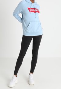 Hellblaue Kapuzenjacke mit rotem Levi's-Logo, verstellbaren Kordeln und Fronttasche; kombiniert mit schwarzen Leggings und weißen Turnschuhen.