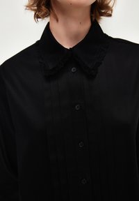 Camicia nera con bottone, dotata di un colletto rifinito in pizzo, caratterizzata da pieghe verticali e cinque bottoni sul davanti per un aspetto strutturato.