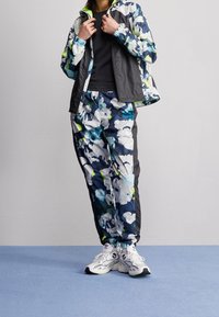 Blomster-mønstret jakke i nuancer af blå, grøn og hvid med en sort bund; matchende blomsterbukser; hvide sneakers med sorte detaljer.