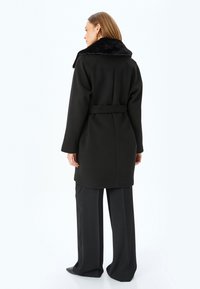 Cappotto in misto lana nero con colletto in pelliccia sintetica, vita con cintura e taglio su misura. Tessuto liscio, design di media lunghezza e senza chiusure visibili.