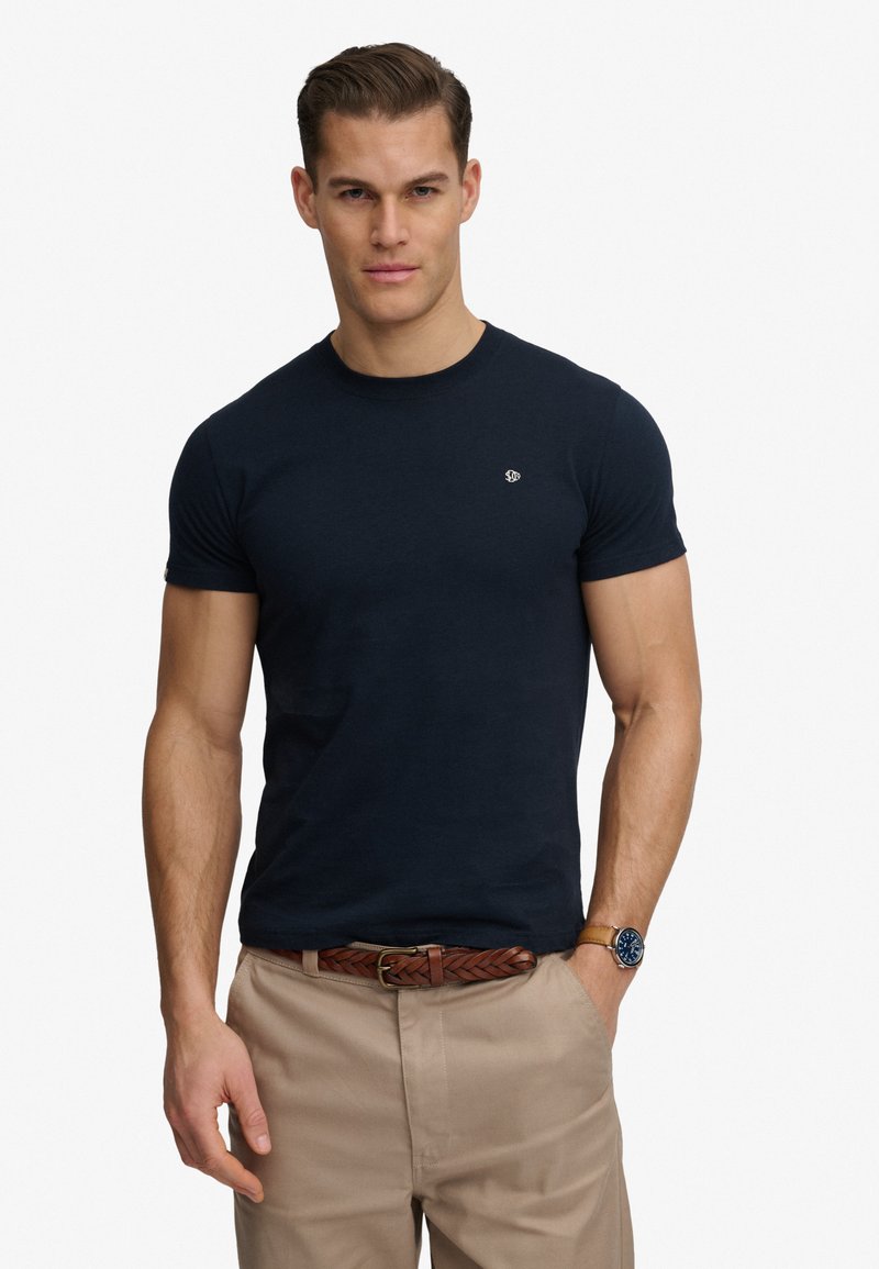 Homme portant un t-shirt noir ajusté, un pantalon beige avec une ceinture marron tressée et une montre-bracelet, posant devant un fond blanc uni.