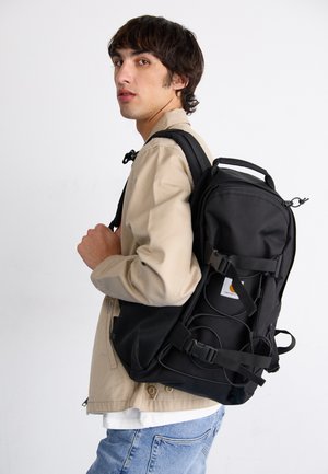 Black backpack with textured fabric, adjustable straps, and a front mesh pocket, worn by a person in a beige jacket and blue jeans.