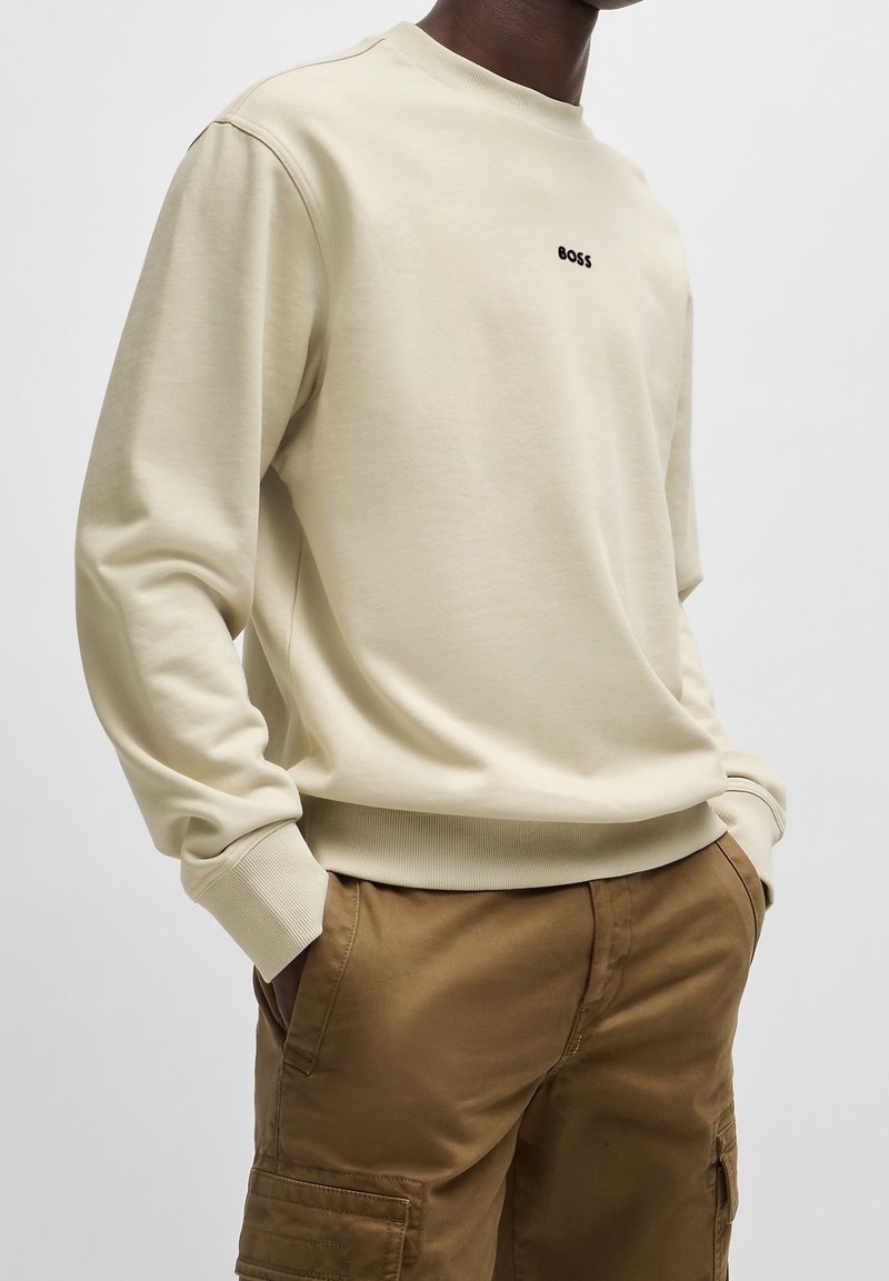 Beige mjuk sweatshirt med rund hals och ribbad mudd. Har en liten svart "BOSS"-logotyp på bröstet. Kombinerad med bruna cargoshorts.