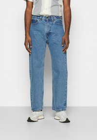Jeans in denim blu chiaro con taglio dritto, caratterizzati da un classico design a cinque tasche e dettagli di cucitura arancione, indossati con sneakers bianche.