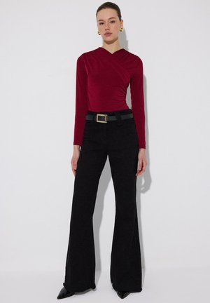 Vrouw draagt een bordeauxrode top met lange mouwen en plooien, gecombineerd met hoge zwarte wijde broek, zwarte riem en zwarte puntige schoenen.