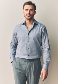 Világoskék kockás ing, hajtott gallérral és gombolós sávval, szürke pliszírozott nadrággal párosítva. Sima anyagtextúra.